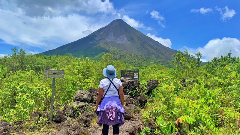This image shows a hiker looking at the iconic, cone-shaped Arenal Volcano (Volcán Arenal). It is located within the Arenal Volcano National Park near the town of La Fortuna in Costa Rica.