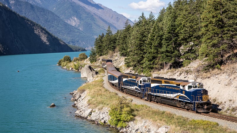 The Rocky Mountaineer, a famous luxury scenic train operating in North America. The specific shot captures the train winding along the turquoise waters of Seton Lake in British Columbia, Canada.