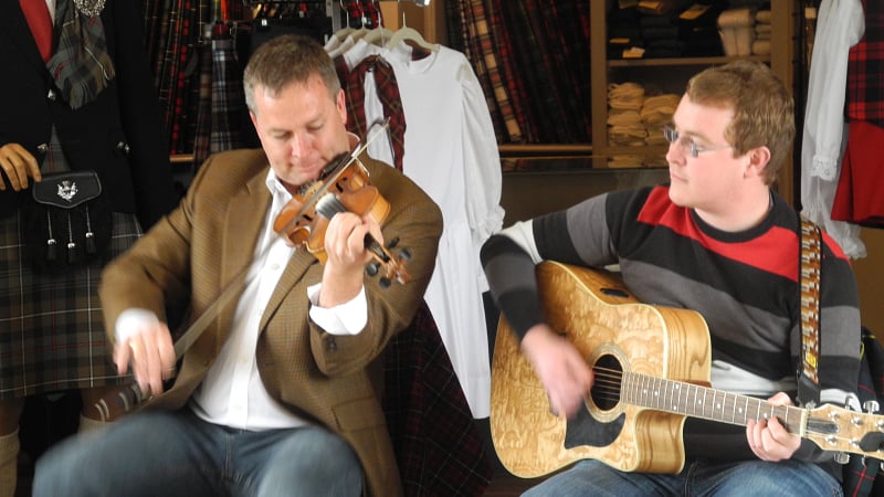 Two musicians participating in a traditional Scottish music session inside a shop filled with Scottish attire, such as kilts and tartans.