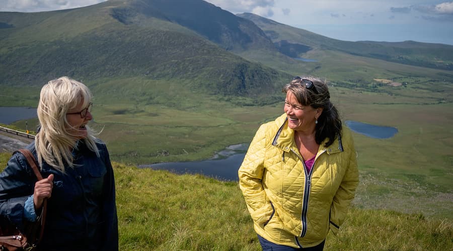 Two adults in jackets standing on a grassy hillside overlooking the Dingle Peninsula, Ireland, with rolling green mountains, winding lakes, and coastal landscape in the background.
