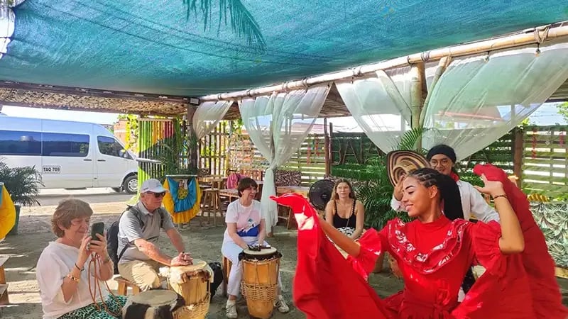 The image captures a tourist group participating in a traditional Afro-Colombian drumming and dance workshop in La Boquilla, a coastal village located just outside of Cartagena, Colombia.