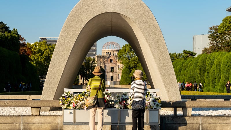 This image shows the Cenotaph for the A-bomb Victims framing the Atomic Bomb Dome in the background, located at the Hiroshima Peace Memorial Park in Hiroshima, Japan.