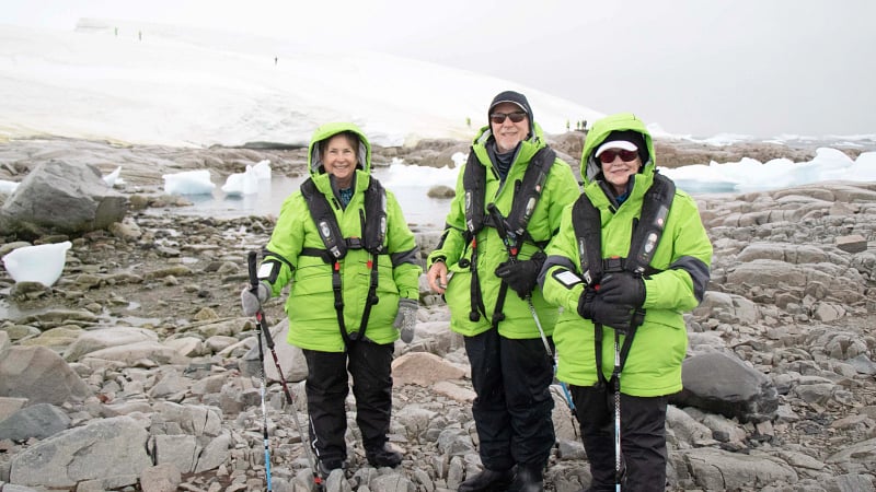 tourists dressed in polar expedition gear standing on a rocky shoreline, during an excursion in Antarctica.