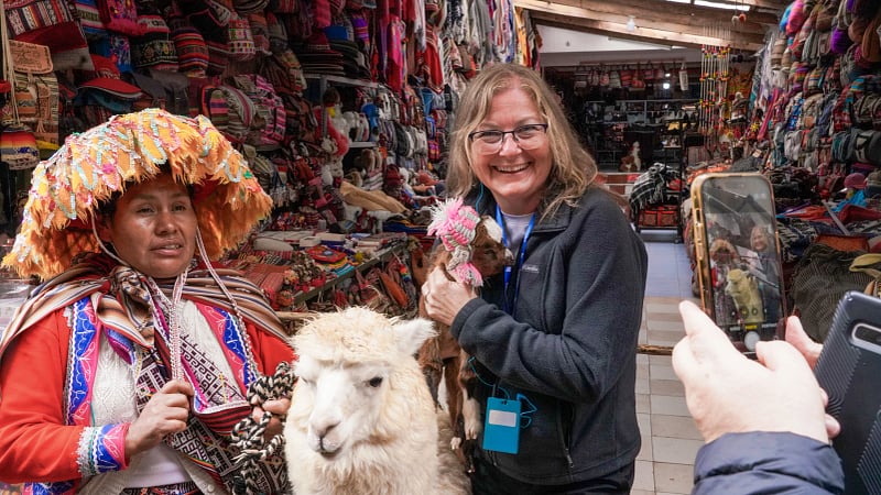 a tourist interacting with a local Quechua woman and domestic alpacas inside a traditional Peruvian artisan textile market.